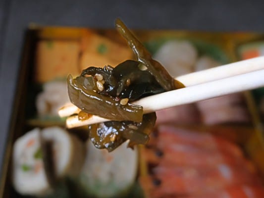 博多久松おせち「博多」弐の重の食材㉓茎わかめの胡麻和えを箸でつまんでいる。２センチほどの長さにカットされた茎ワカメにゴマがついている。あっさりとした見た目