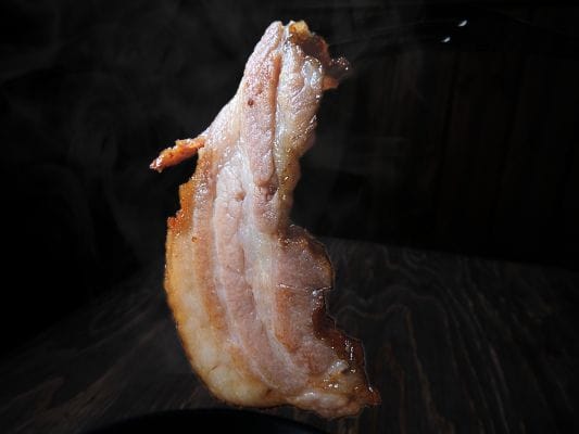 グリルした厚さ約7mmのベーコンをフォークで刺している。グリルしたてで湯気が立っている。